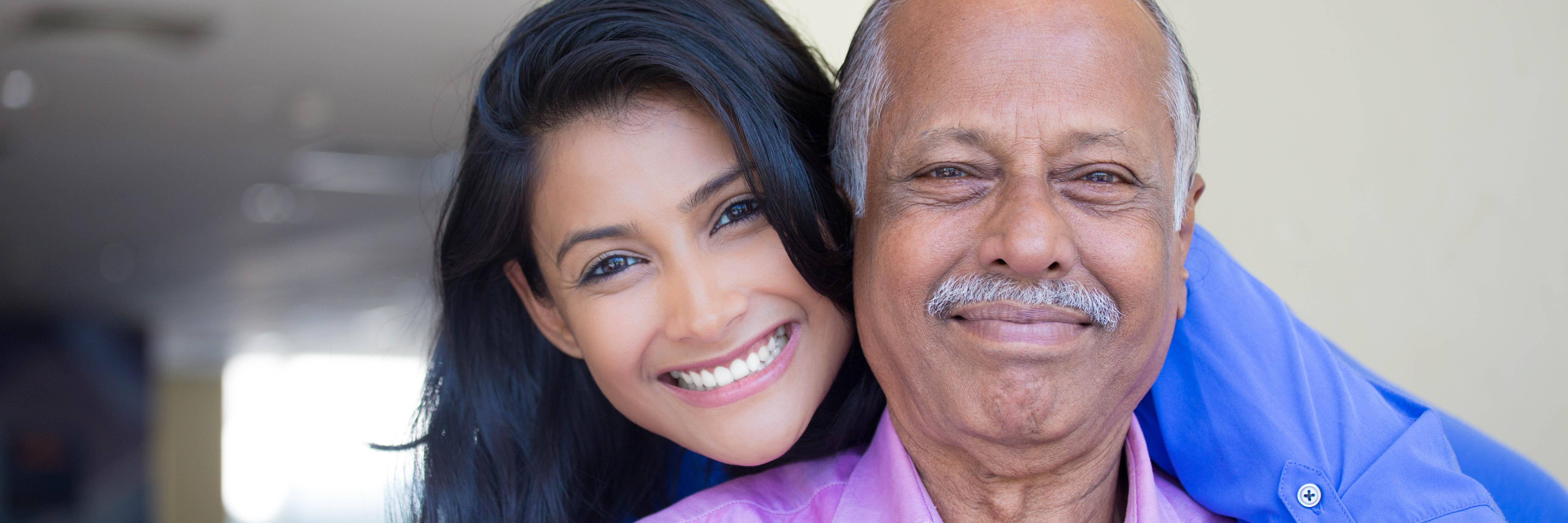 Close-up portrait of a young woman in a blue shirt holding older man in pink collar indoors.